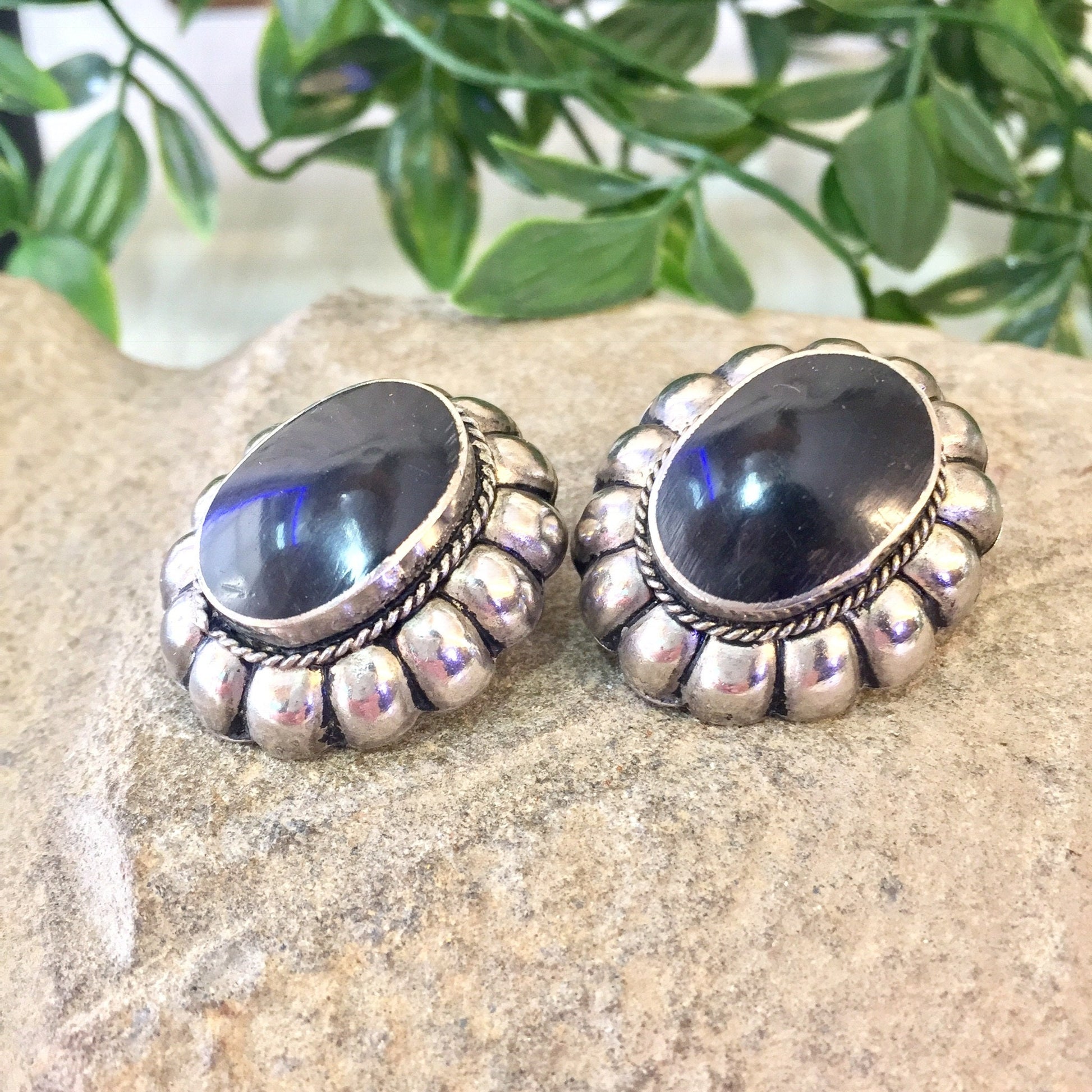 Vintage sterling silver clip-on earrings with black onyx stones in a Southwestern style, resting on a stone surface with greenery in the background.