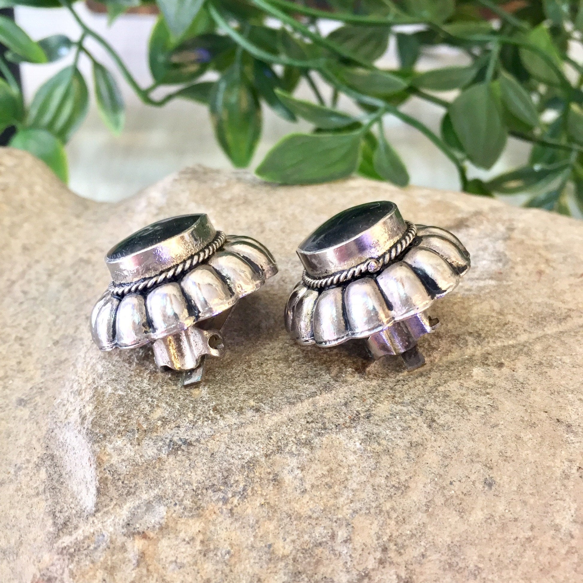 Vintage sterling silver clip-on earrings with black onyx in a Southwestern style, displayed on a stone surface with greenery in the background.