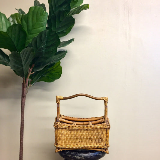 Vintage wicker utensil caddy with handle next to a green plant against a plain background.