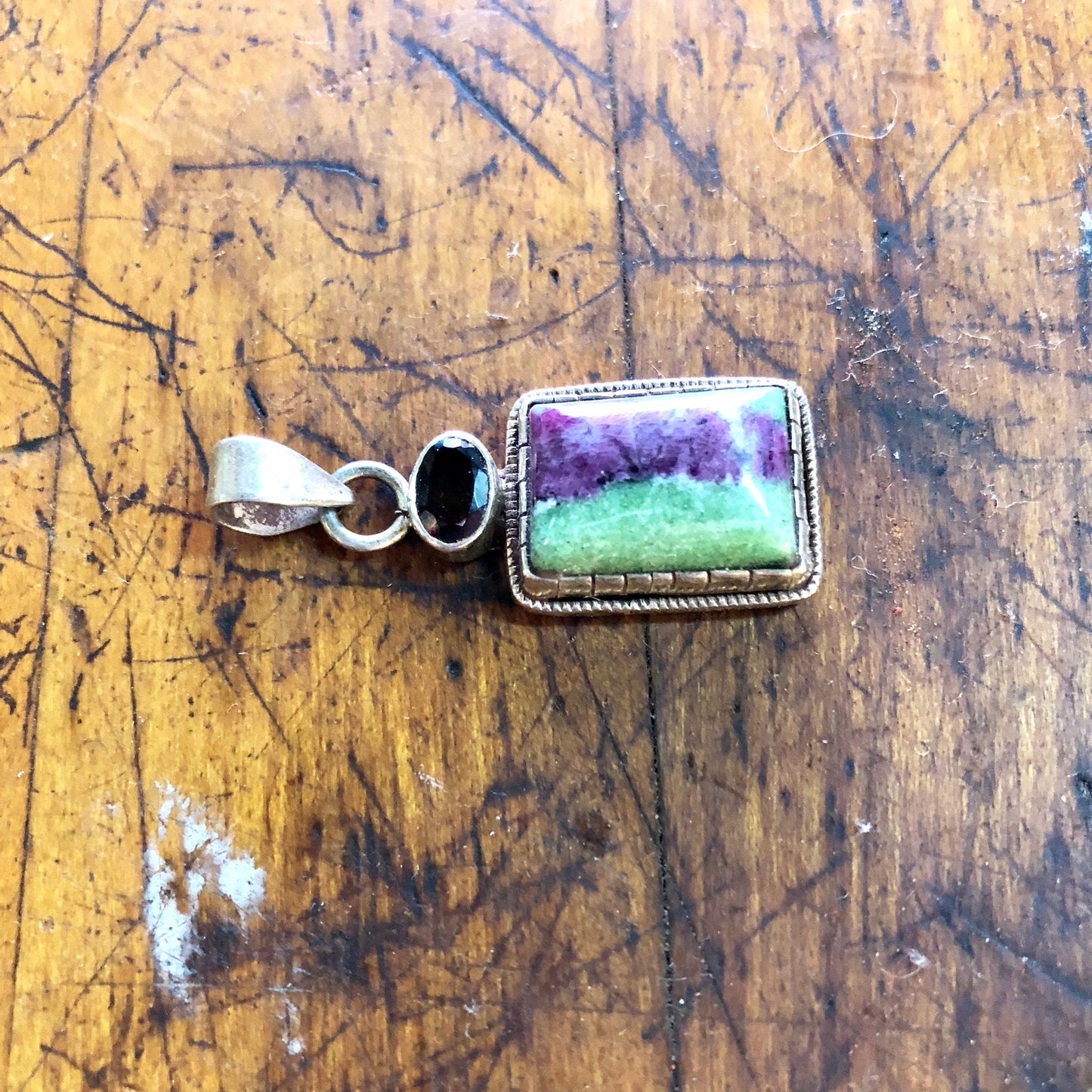 Vintage sterling silver pendant with green and purple natural stone, possibly malachite or jade, accented with a red garnet gemstone, against a weathered wooden background.