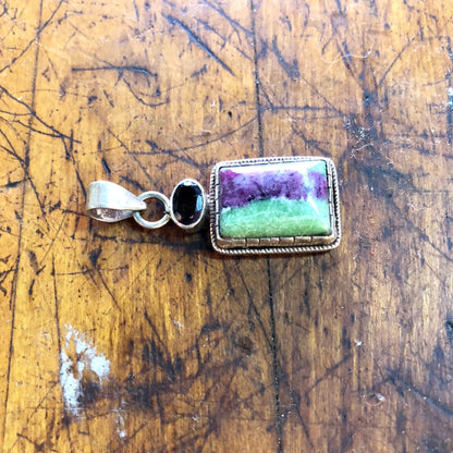 Vintage sterling silver pendant with green and purple natural stone, possibly malachite or jade, accented with a red garnet gemstone, against a weathered wooden background.