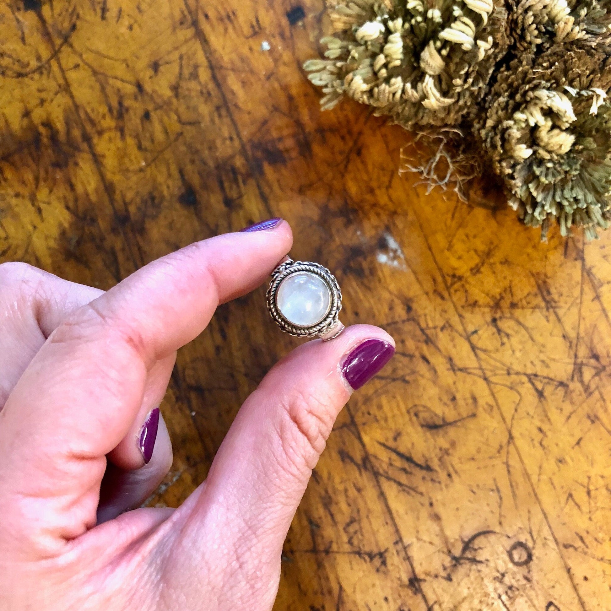 Vintage sterling silver ring with iridescent white moonstone cabochon, held in hand over wooden surface with decorative pine cones