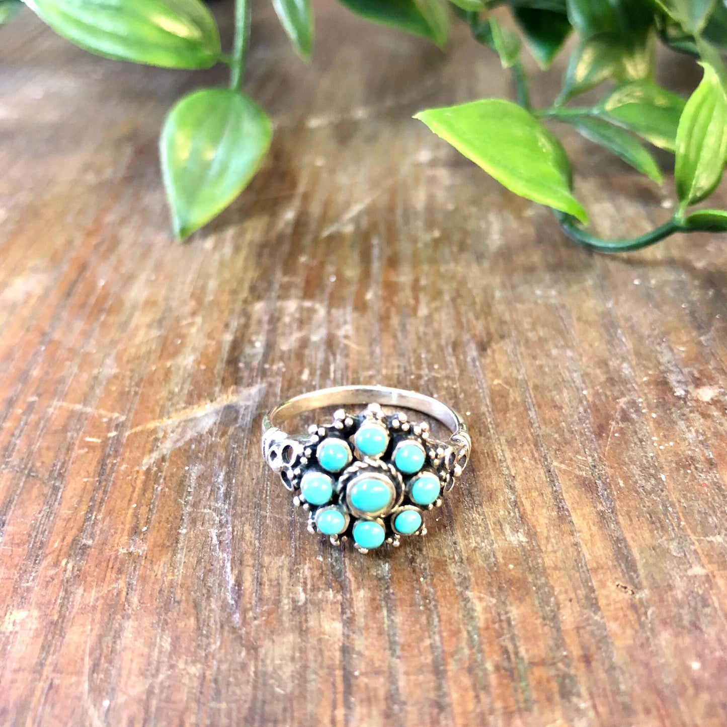 Vintage sterling silver ring featuring a flower-shaped cluster of turquoise beads set against a wooden surface with green leaves in the background