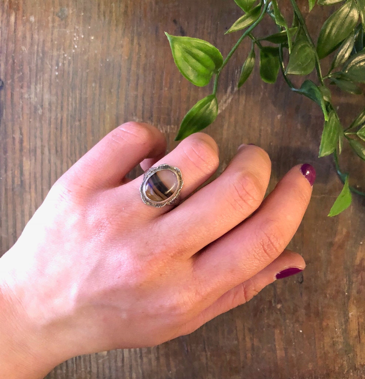 Vintage sterling silver ring with brown striped agate stone on hand next to green plant leaves on wooden table