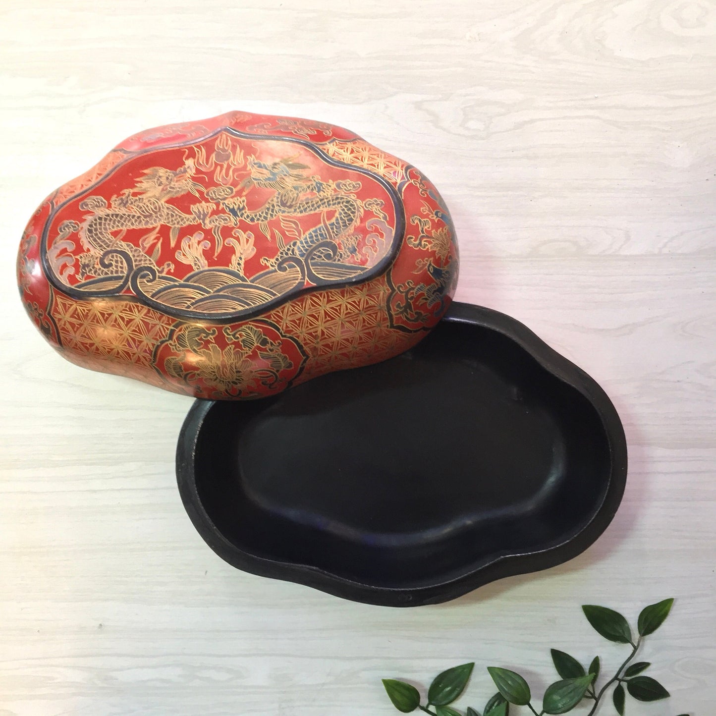 Vintage Asian red laminated box with gold dragon design, accompanied by a black catch-all dish, surrounded by green leaves on a white wooden surface.