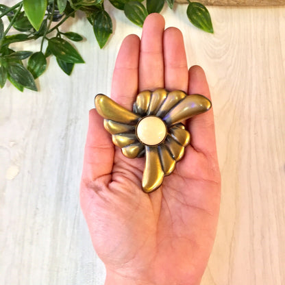 Hand holding a large vintage gold-tone flower clip-on earring with a white center against a light wood background with green foliage.