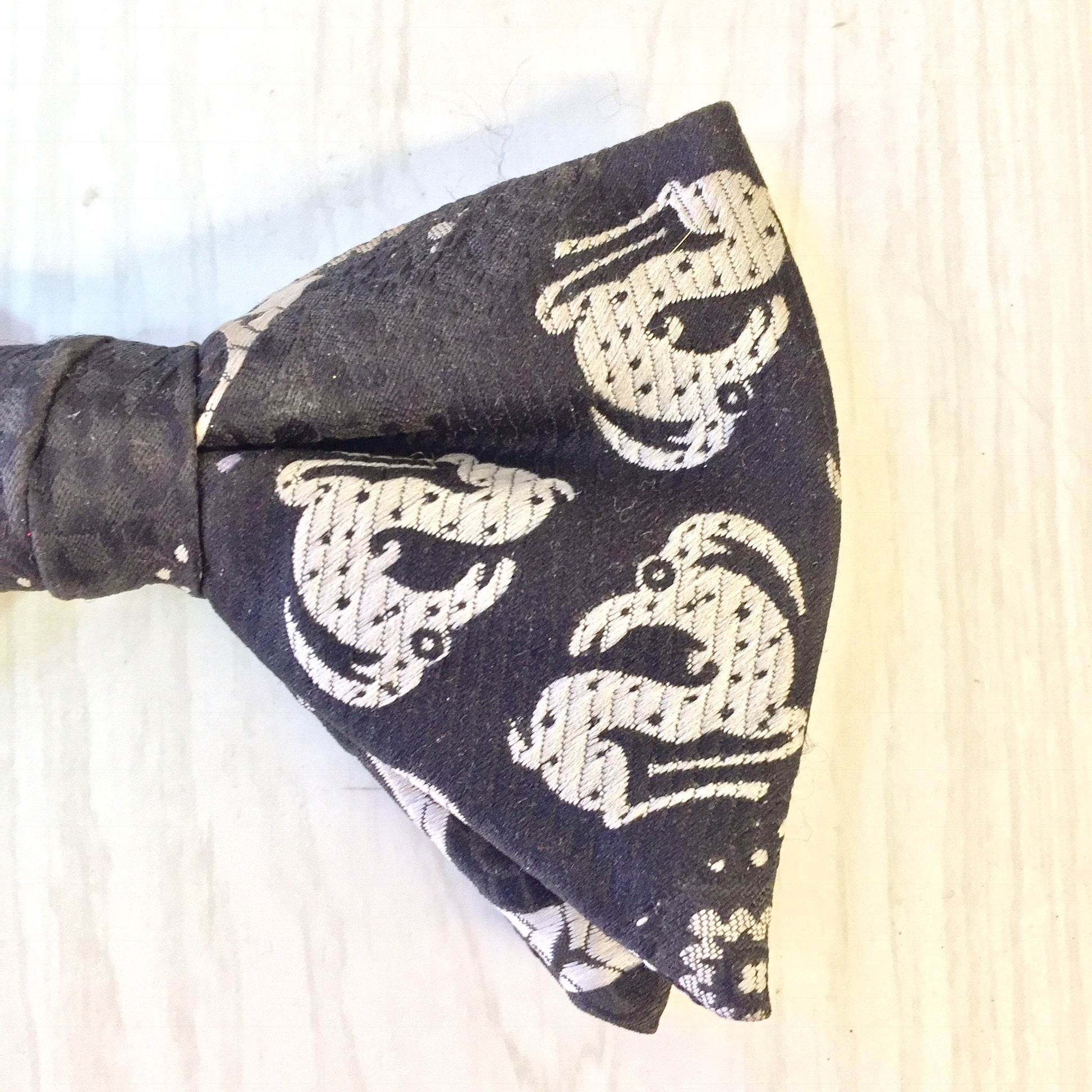 Black bow tie with white floral pattern, clipped on partial white shirt collar, on light wooden background.