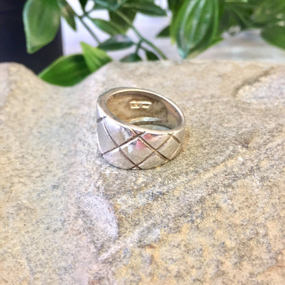 Vintage sterling silver statement ring with thick band and criss-cross diamond pattern design, displayed on stone surface with greenery in background.