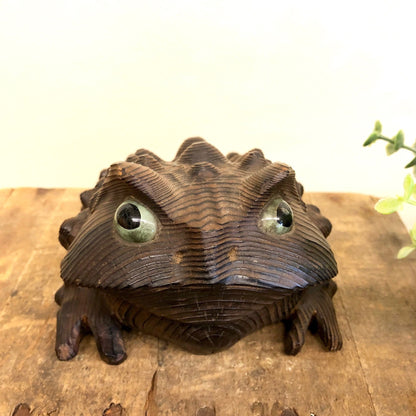 Hand carved wooden frog with textured brown finish and green glass eyes, sitting on wooden surface with flowers in background, vintage animal home decor sculpture.