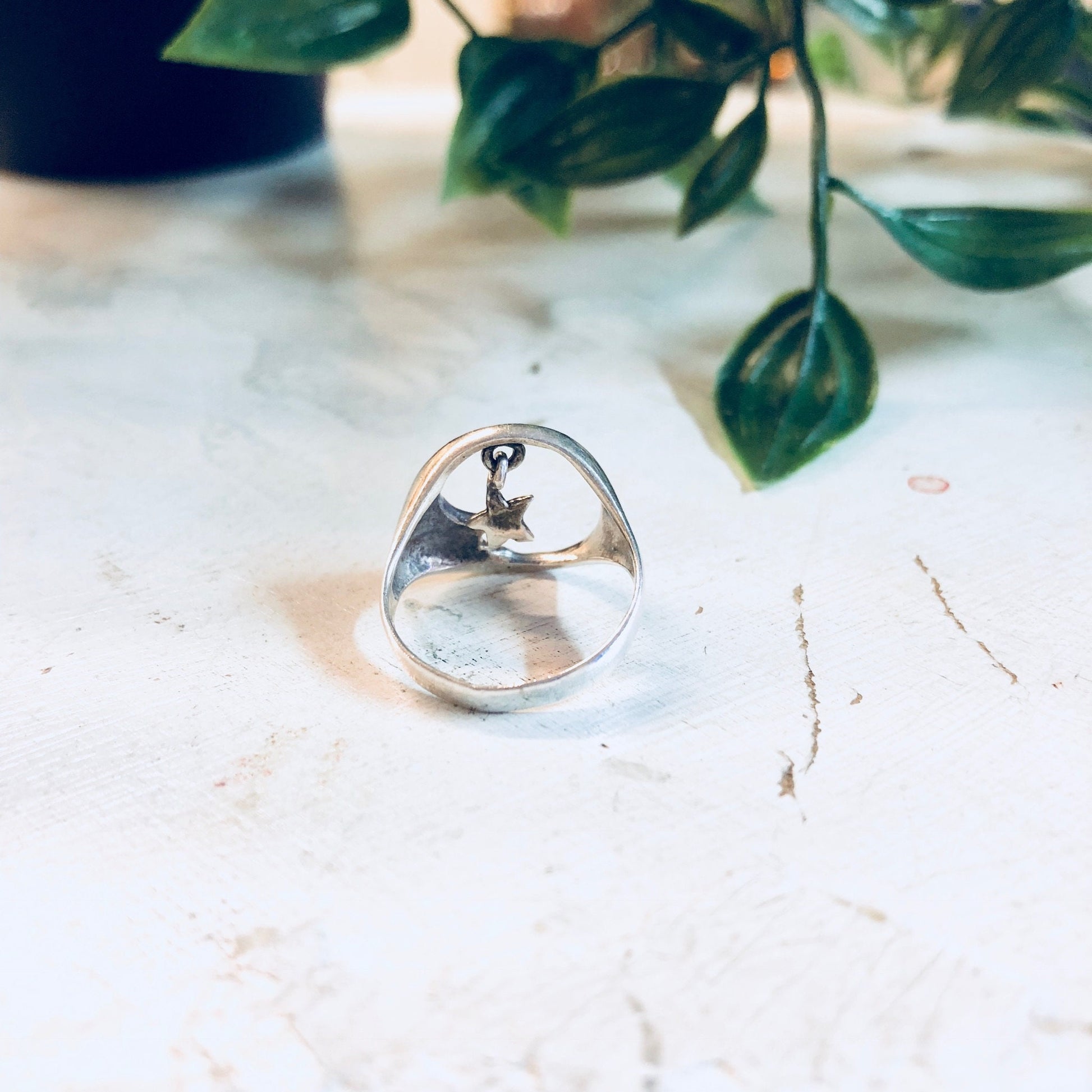 Vintage sterling silver ring with crescent moon and star cutout design, displayed on marble surface with greenery in soft focus background, bohemian style promise ring or statement jewelry piece.