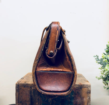 Vintage brown leather handbag with rustic bohemian style from the 1960s, sitting on a wooden surface with a plant in the background.