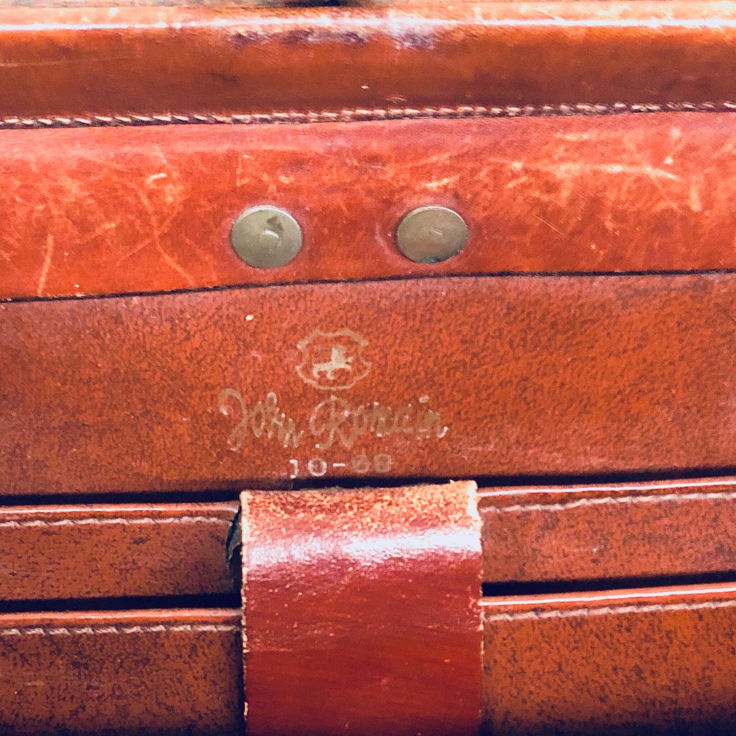 Vintage brown leather John Romain handbag with rustic, bohemian 1960s style and embossed logo on the front, closeup detail of the weathered leather texture and brass rivets.