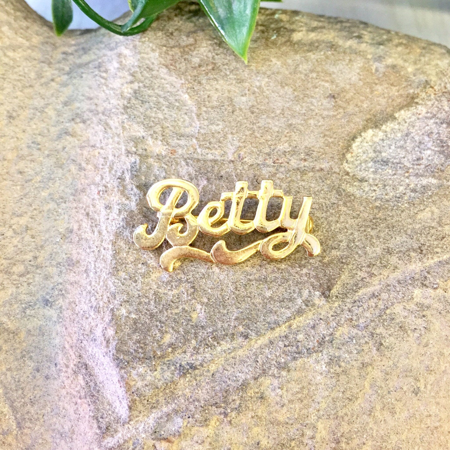 Vintage gold-toned "Betty" name brooch pin resting on textured stone surface with green plant leaves in background