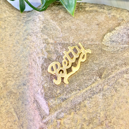 Vintage gold-toned "Betty" name brooch pin lying on textured stone surface with green leaves