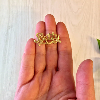 A hand holding a vintage gold-toned "Betty" name brooch against a wooden background.