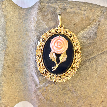 Vintage gold-toned oval pendant featuring a pink rose cameo on a black background, surrounded by intricate gold floral detailing, displayed on a stone surface.