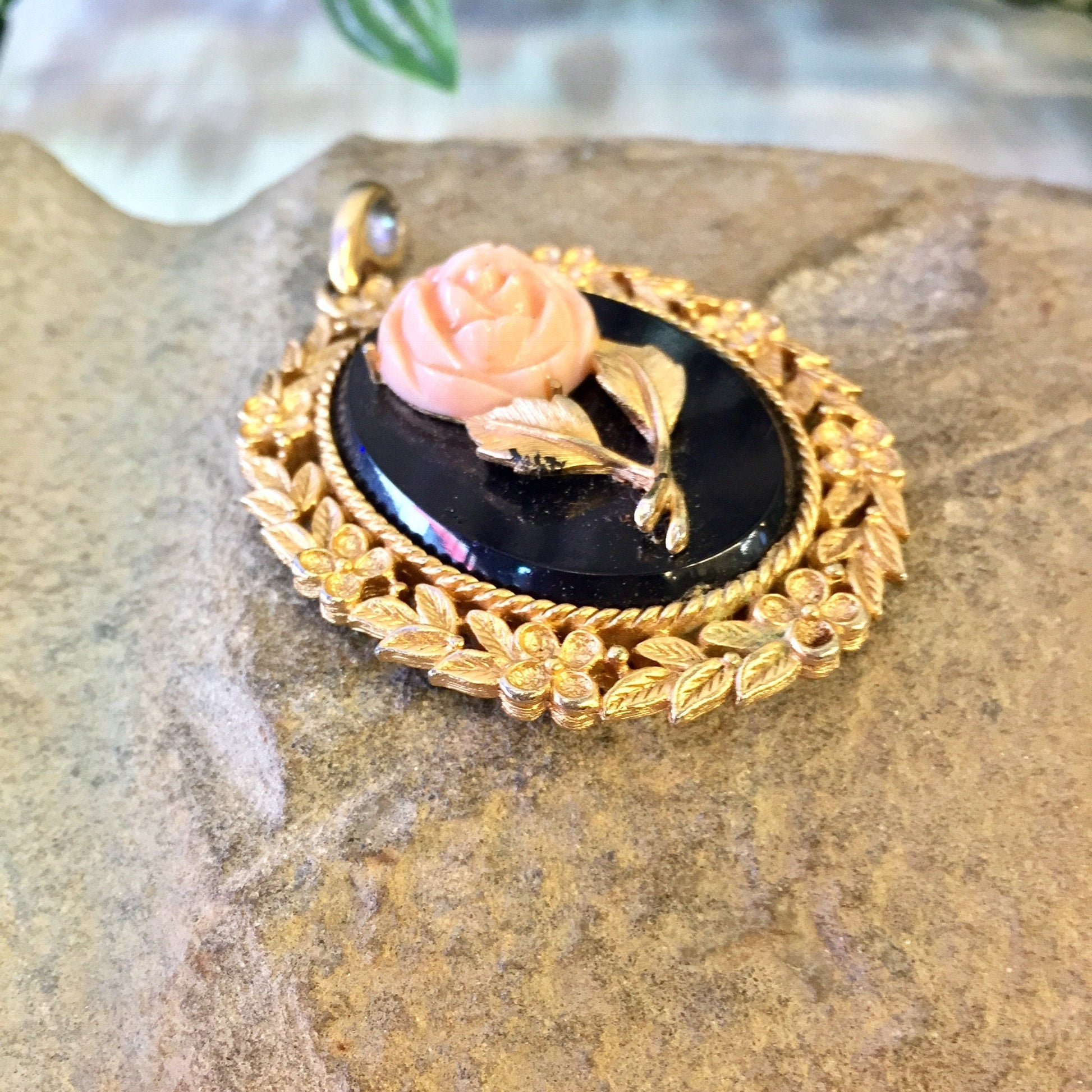 Vintage gold-toned Avon cameo pendant featuring a pink rose on a black background, set on a decorative oval frame with leaf designs, displayed on a textured stone surface.