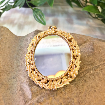 Vintage gold-toned cameo pendant featuring a small mirror surrounded by an intricate floral rose design, set against a rustic stone background with greenery.