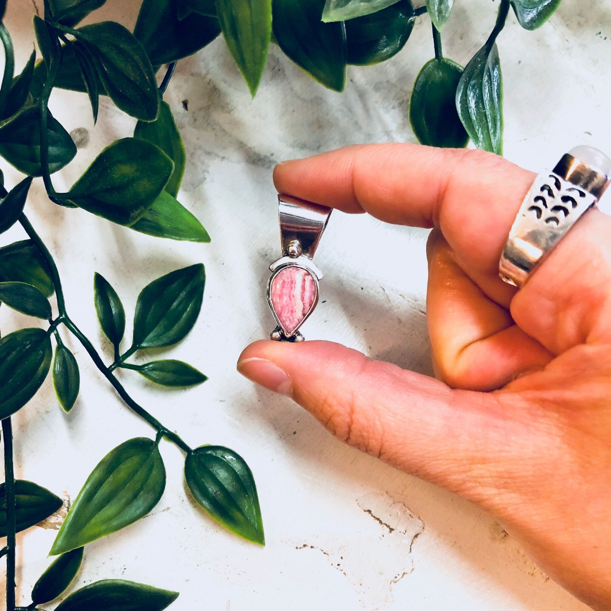 Vintage silver and pink rhodochrosite pendant necklace held in hand against white marble and green foliage background