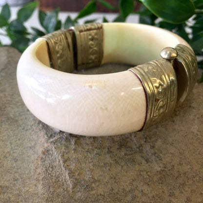 Vintage white and silver hinged bangle bracelet with engraved designs and pin closure, displayed on stone surface with greenery in background.