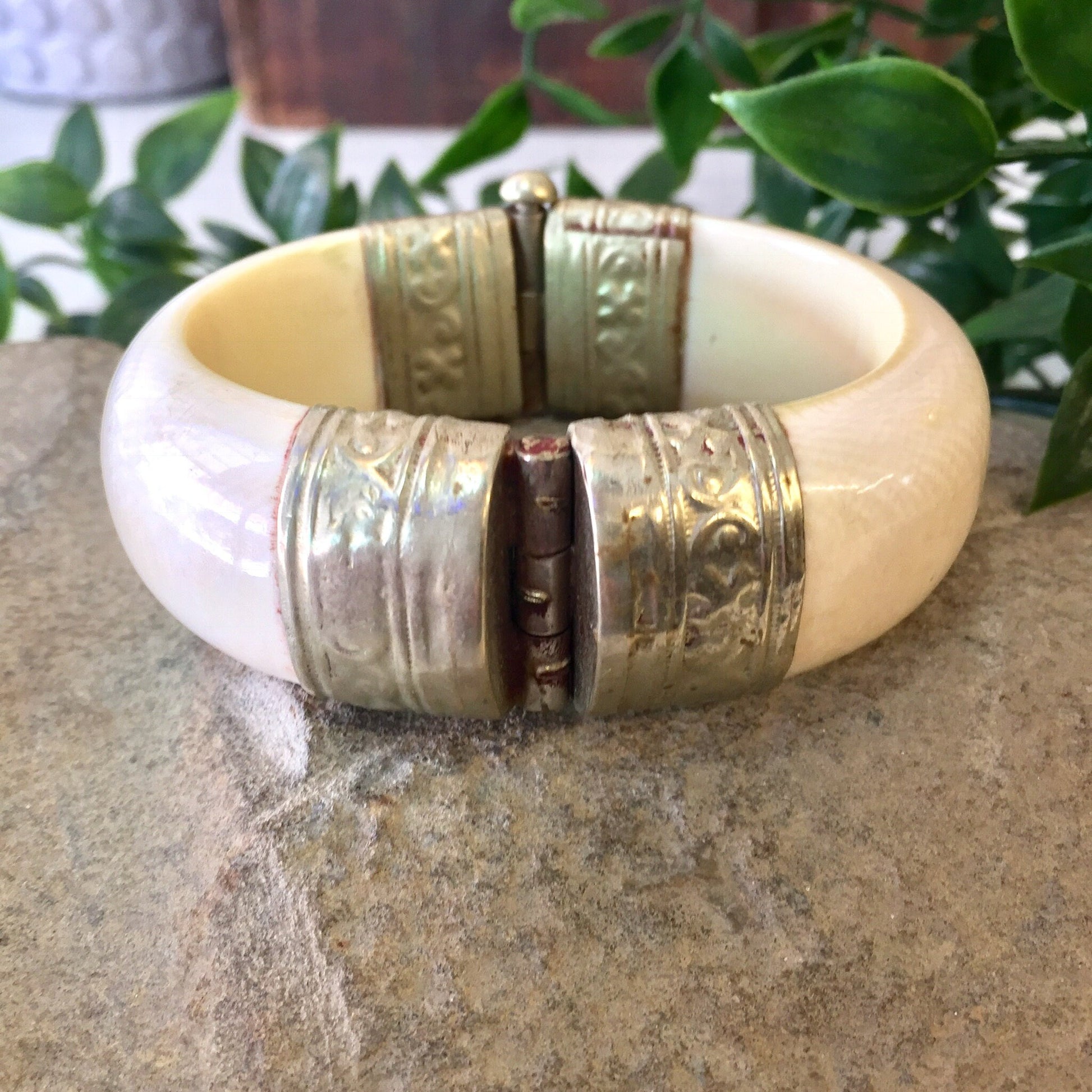 Vintage white and silver hinged bangle bracelet with engraved designs and pin closure, displayed on stone surface with greenery in background.