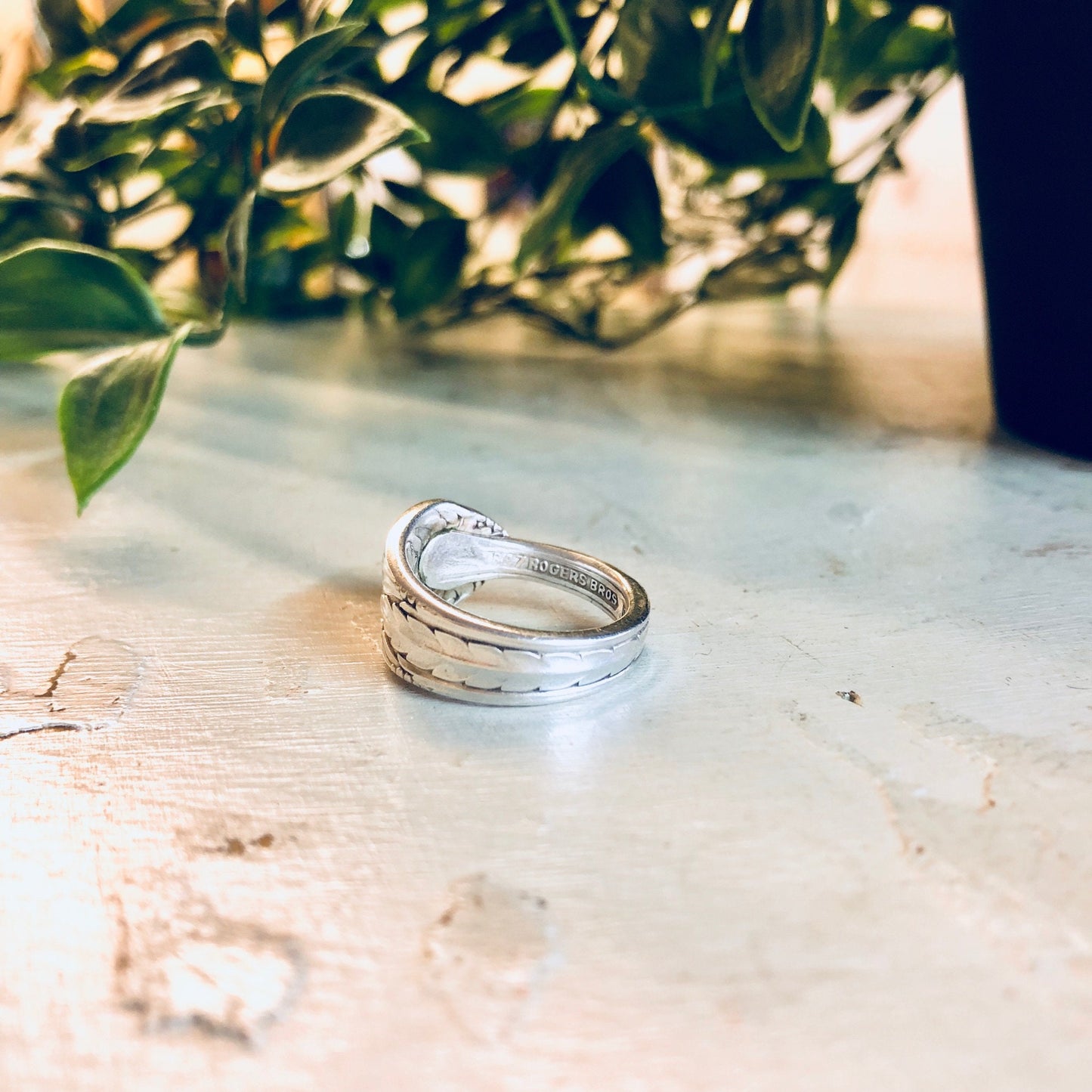 Vintage silver spoon ring with floral design, handcrafted from Rogers Bros silverware cutlery, placed on rustic wooden surface with green foliage in background, unique statement jewelry piece.