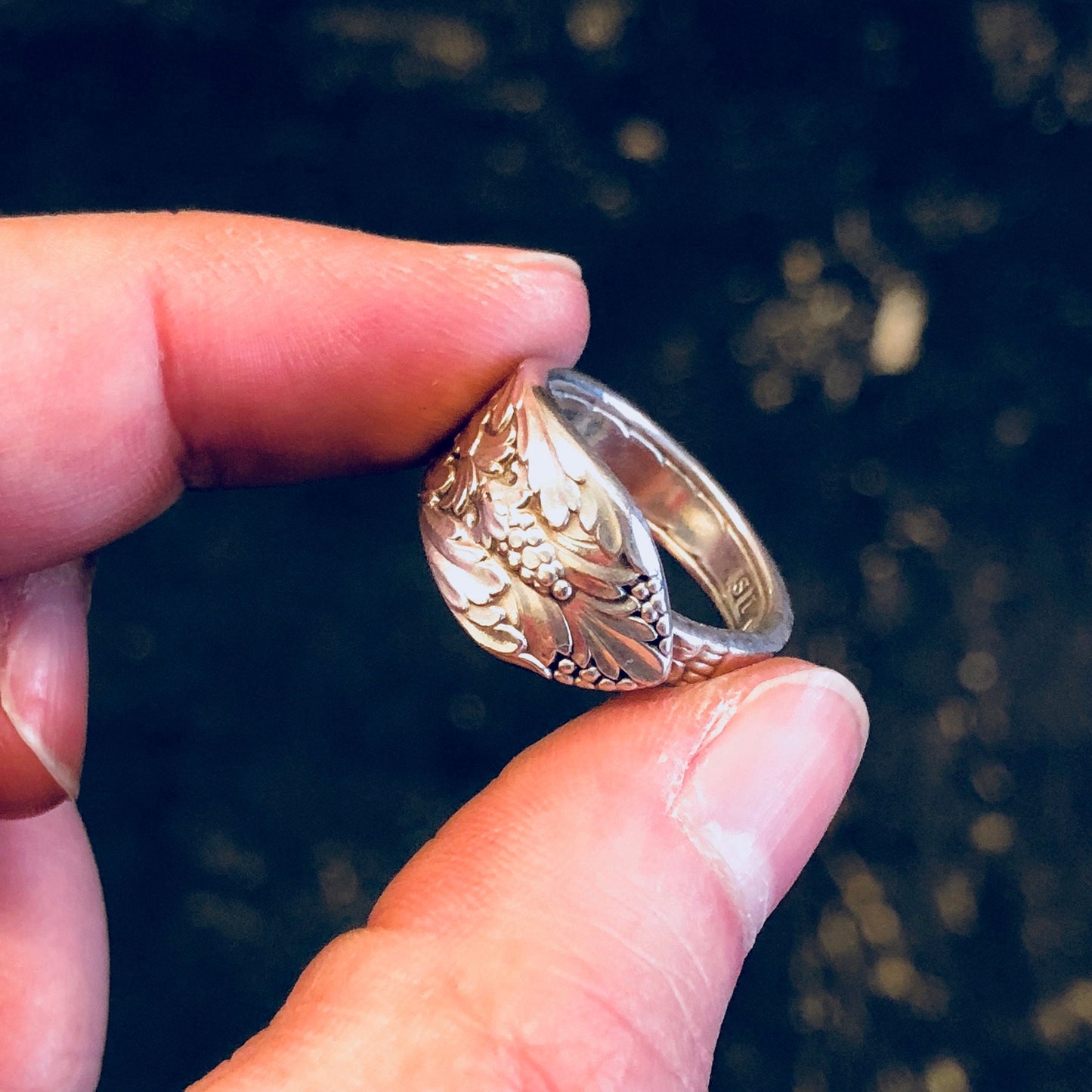 Vintage silver spoon ring with intricate floral design, crafted from Rogers Bros silverware, held between fingers against dark background.