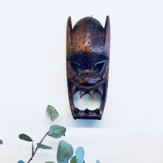 Vintage hand-carved wooden mask with intricate details and dark brown finish, displayed on white wall with green foliage, suitable for primitive home decor or wall hanging.