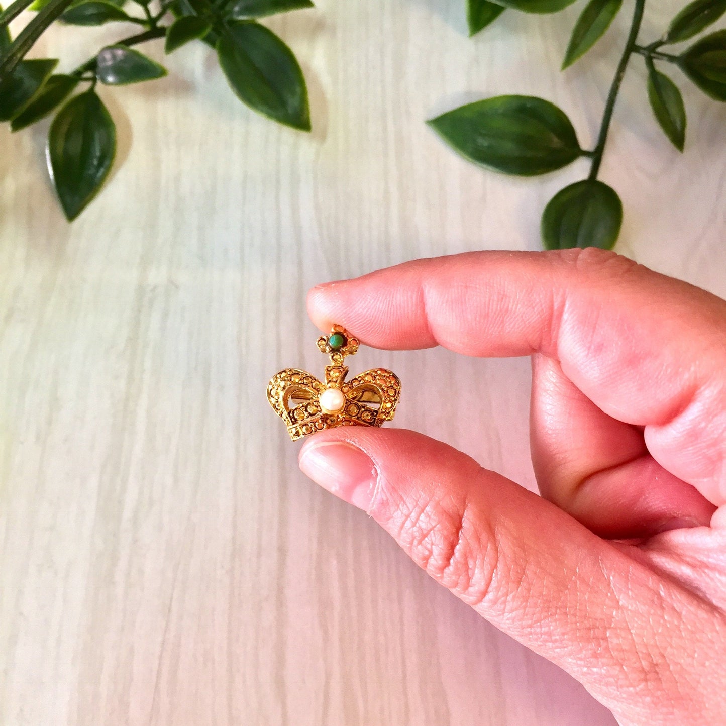 Vintage gold crown brooch pin held in hand near green leaves on wooden surface