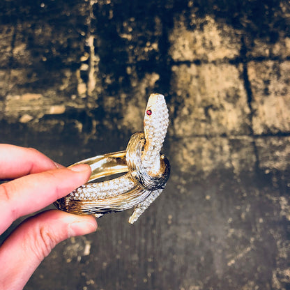 Vintage gold-toned hinged snake bracelet with rhinestones, held in hand against weathered wooden background.