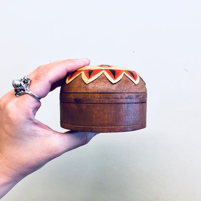 Hand holding a round wooden jewelry box with a carved sun design in red, orange and yellow colors on the lid against a plain background.