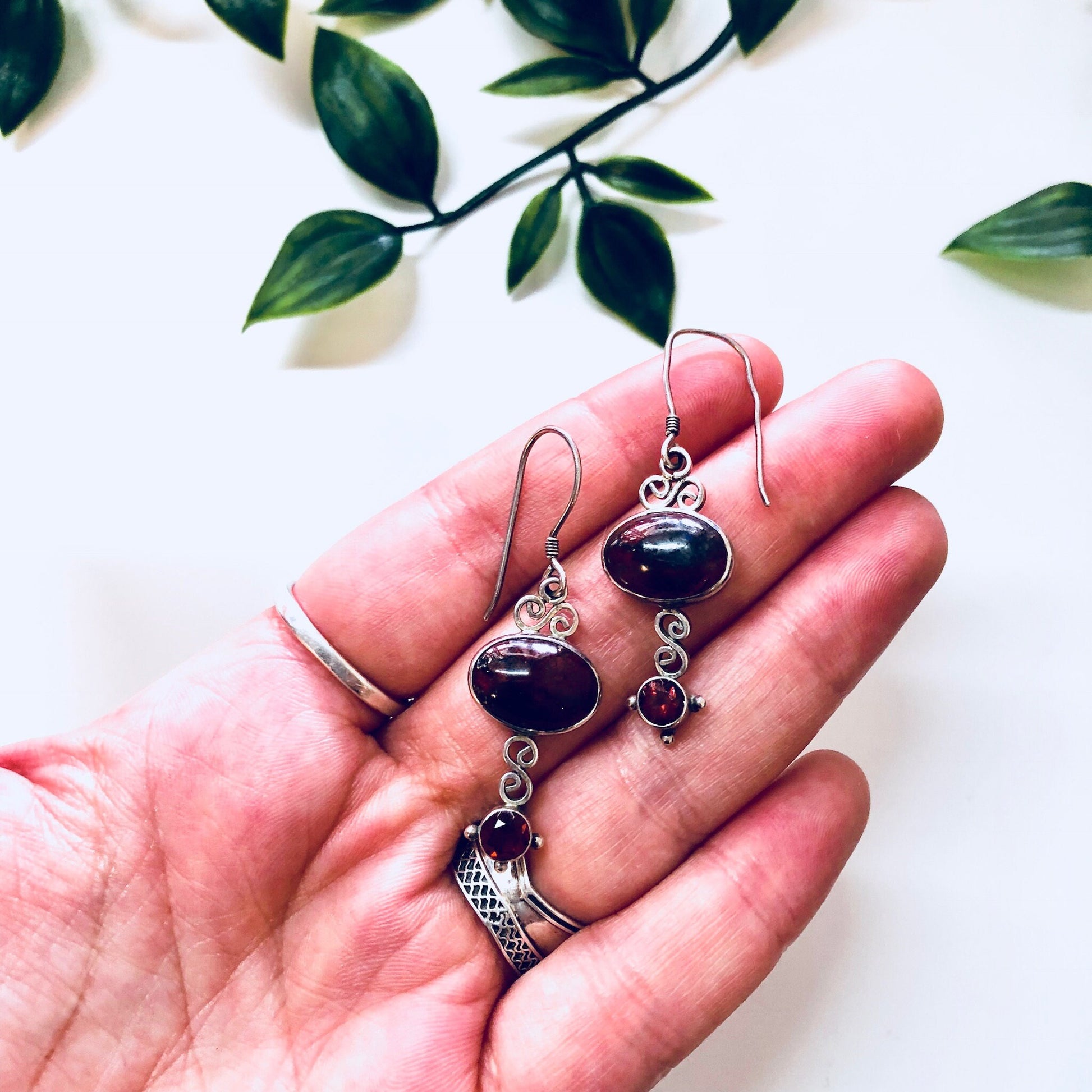 Vintage silver dangle earrings with oval red gemstones, intricate metalwork, and green leafy background