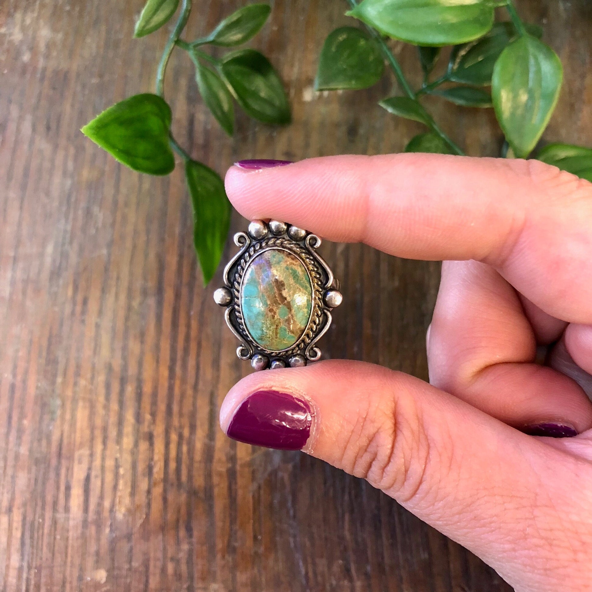 Vintage silver ring with blue Royston turquoise cabochon stone set in ornate sterling silver mounting, held between fingers with dark red nail polish against a rustic wooden background with green plant leaves.