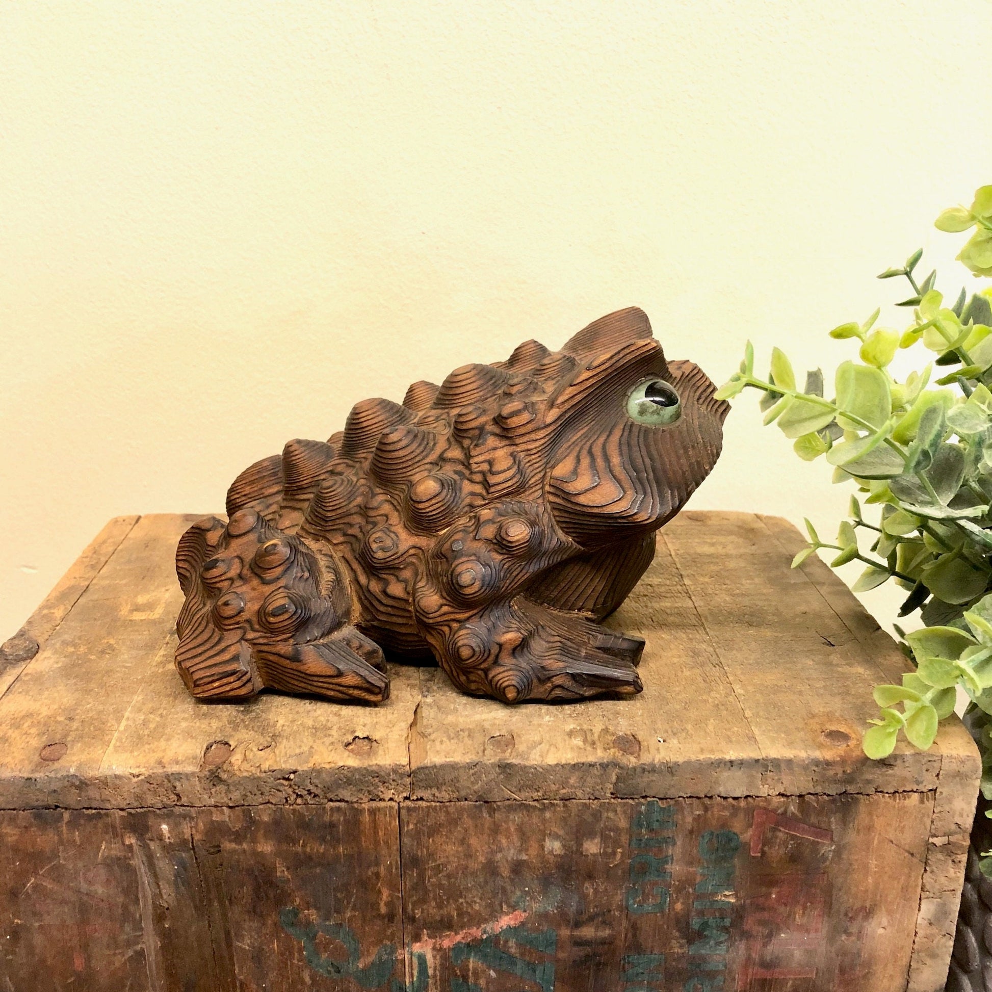 Vintage hand carved wooden frog figurine sculpture for rustic animal home decor, sitting on distressed wooden surface with greenery in background