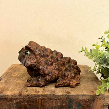 Intricately carved wooden frog figurine with textured skin, sitting on weathered wood surface beside green foliage, providing vintage animal-themed home decor in brown and black tones.