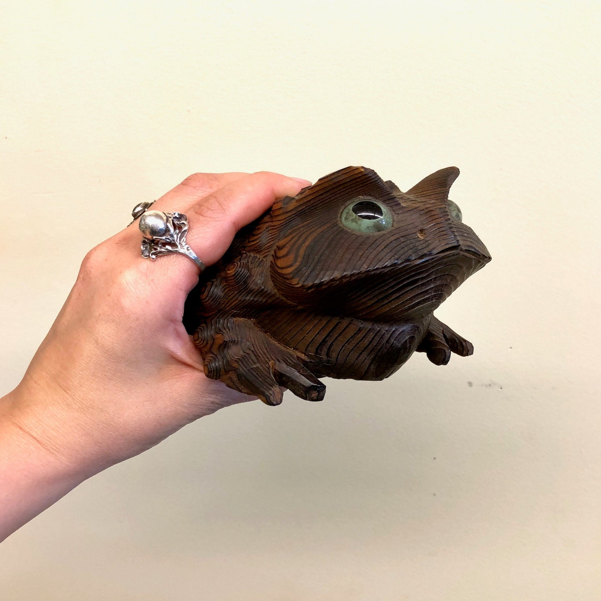 Hand holding a vintage carved wooden frog figurine with brown, black and green tones, suitable for home decor or mantlepiece display.