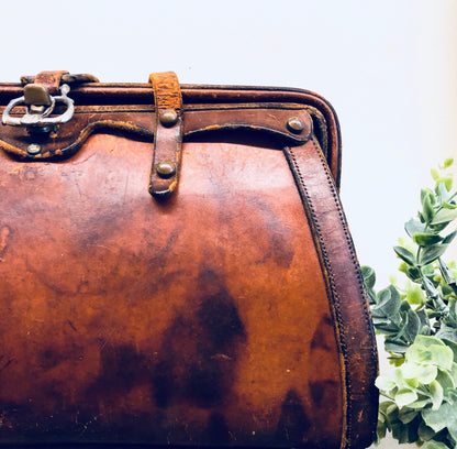 Vintage brown leather handbag with rustic, bohemian style from the 1960s era, featuring a distressed patina and metal clasp closure, photographed with greenery accent.