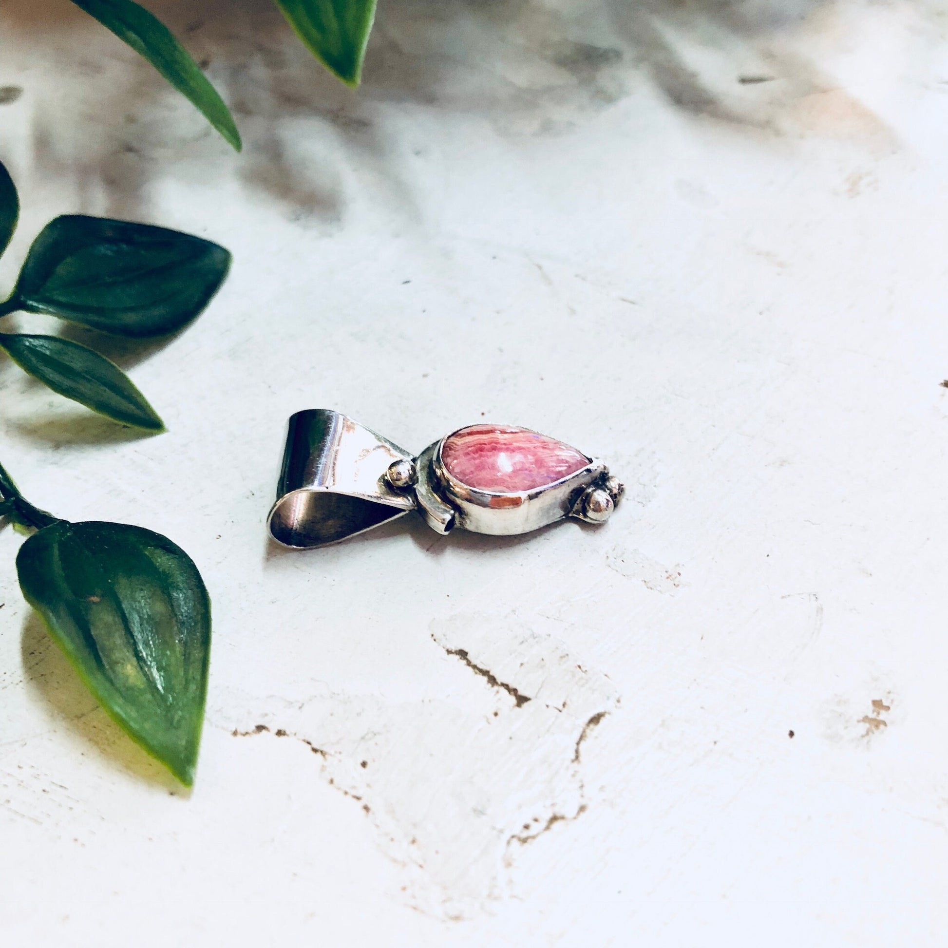 Vintage sterling silver pendant with pink rhodochrosite gemstone, surrounded by lush green foliage on a white marble surface.