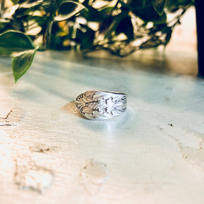 Vintage silver spoon ring with floral design, sitting on rustic wooden surface with green foliage in background, handmade cutlery jewelry statement piece by Rogers Bros.