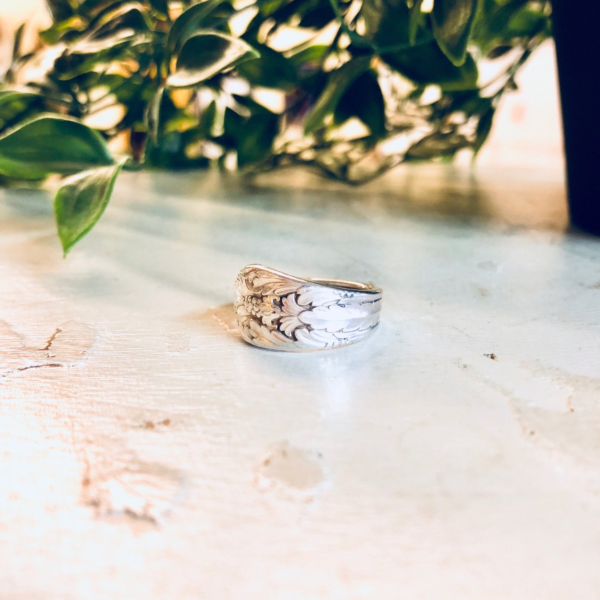 Vintage silver spoon ring with floral design on band, handcrafted from Rogers Bros silverware cutlery, placed on weathered concrete surface with greenery in soft focus background.