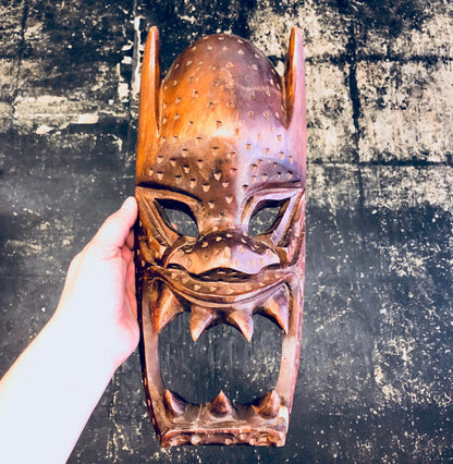 Intricately carved wooden mask with spiked ears and teeth, held in a hand against a grungy textured background.