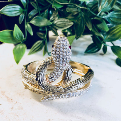 Vintage gold-toned hinged snake bracelet with rhinestones, displayed on stone surface with green leafy background.