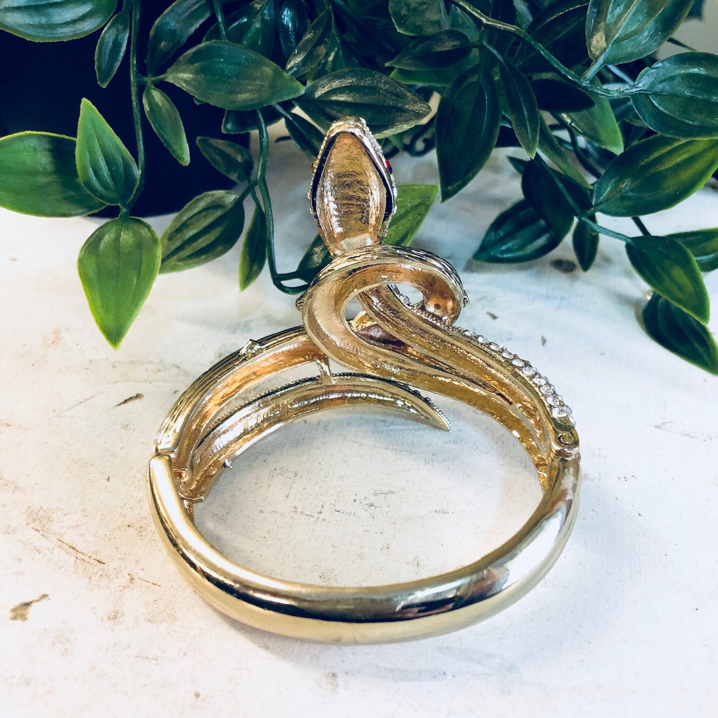 Vintage gold-toned hinged snake bracelet with rhinestones, displayed on a white surface with green foliage in the background.