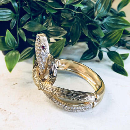 Vintage gold-toned snake bracelet with rhinestone accents and hinged cuff design, displayed on white surface with leafy green foliage background.