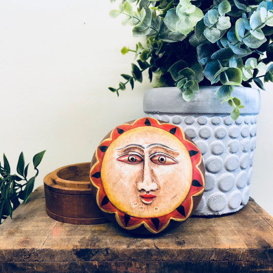 Wooden sun box with carved face and red, orange and yellow colors next to a potted plant on a wooden surface.