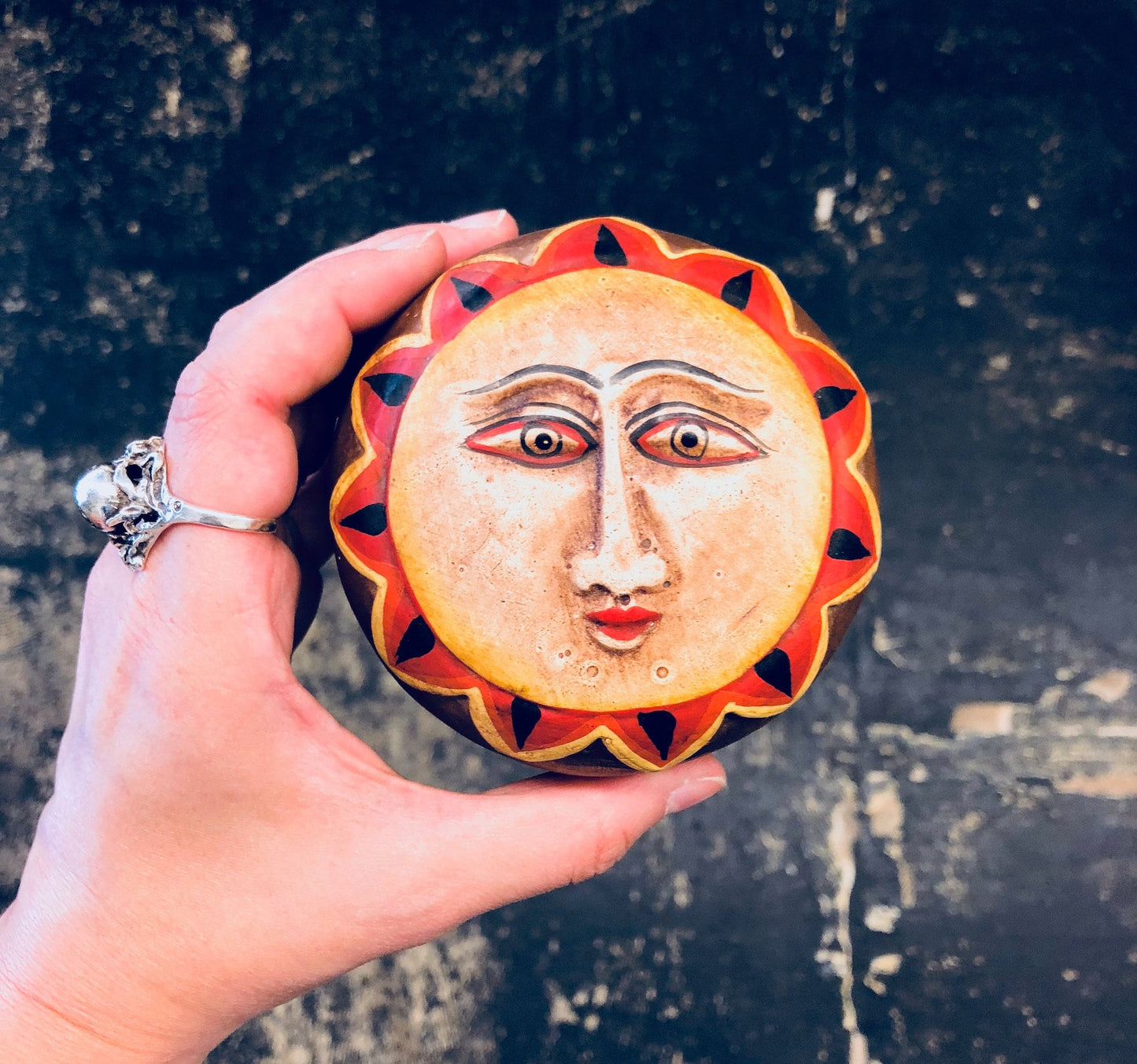Wooden sun face trinket box held in hand, with red, orange and yellow carved rays surrounding facial features on a textured dark background.