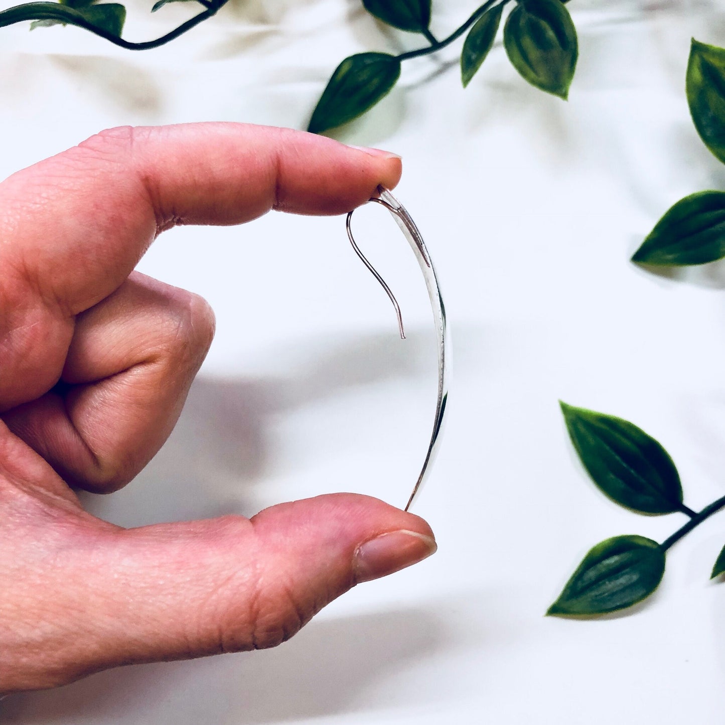 The image shows a hand holding up a simple, curved silver wire earring against a white fabric background with green leaves. The earring appears to be made of thin, polished sterling silver wire in a minimalist, abstract design. It is a unique vintage or handmade piece of statement jewelry that could make a thoughtful anniversary gift.