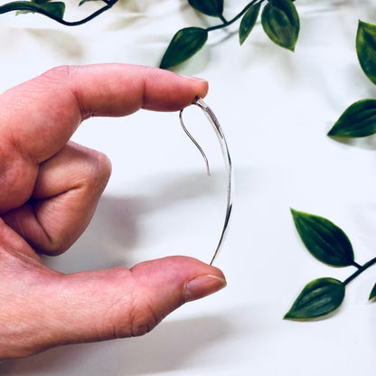 The image shows a hand holding up a simple, curved silver wire earring against a white fabric background with green leaves. The earring appears to be made of thin, polished sterling silver wire in a minimalist, abstract design. It is a unique vintage or handmade piece of statement jewelry that could make a thoughtful anniversary gift.