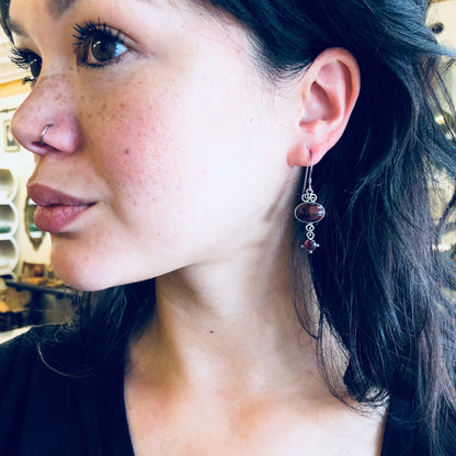 Close-up of a woman wearing vintage silver dangle earrings with red gemstones. The earrings have an intricate design and hang from pierced ears. The woman has freckled skin and long dark hair.