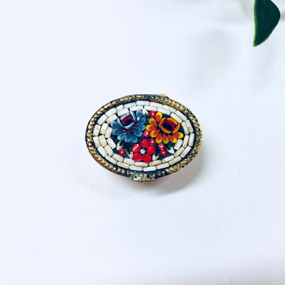 Vintage millefiori mosaic pill box with gold trim featuring red, blue, orange and white swirl design on a white background with a green leaf.